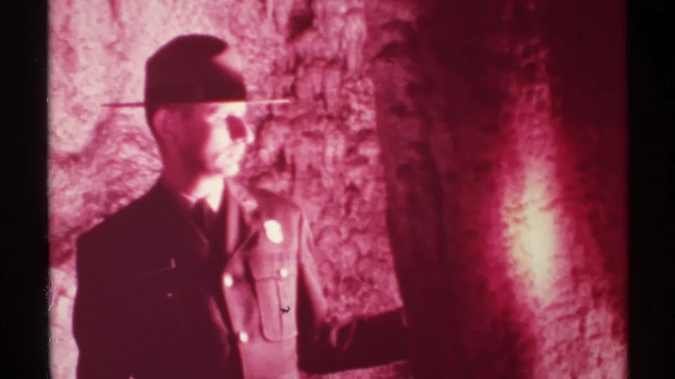 Adult man in a cap and uniform-like shirt raises one arm beside the red cave wall in 1972 New Mexico home-movie footage.