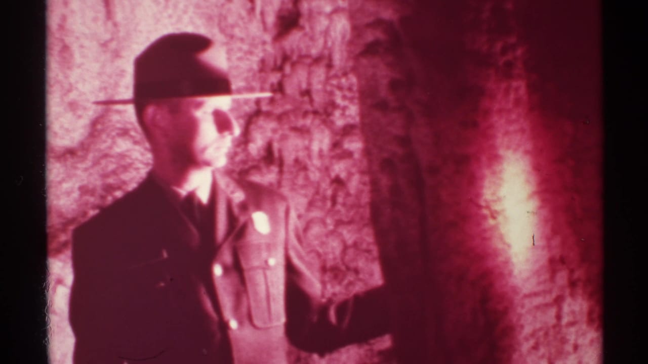 Uniformed man in a wide-brimmed hat holds a flashlight beside rough cave walls.