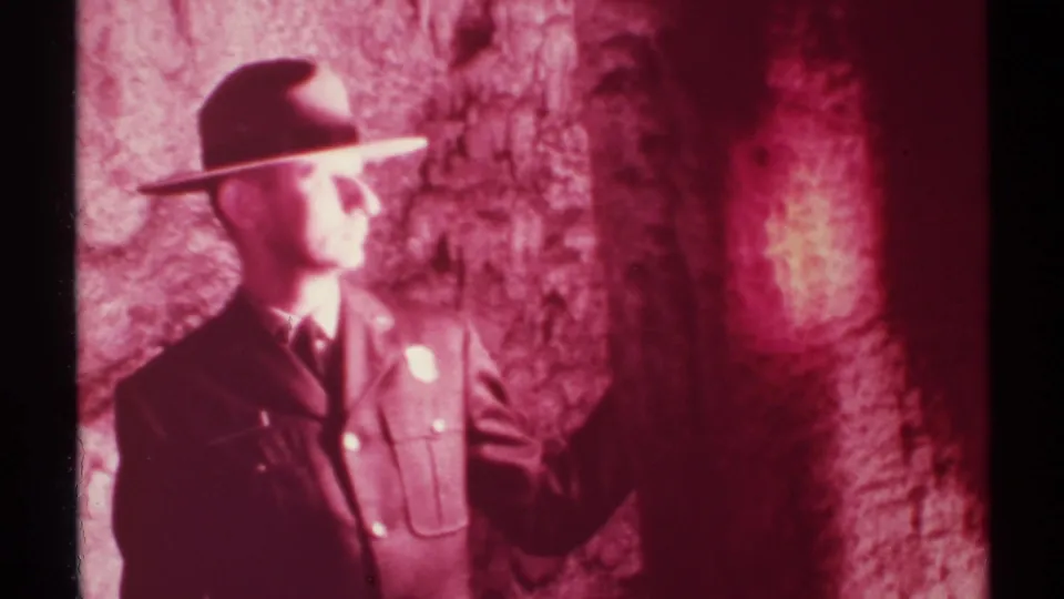 1972 New Mexico 8mm frame showing a uniformed man in a wide-brimmed hat looking toward reddish cave rock while holding a flashlight.