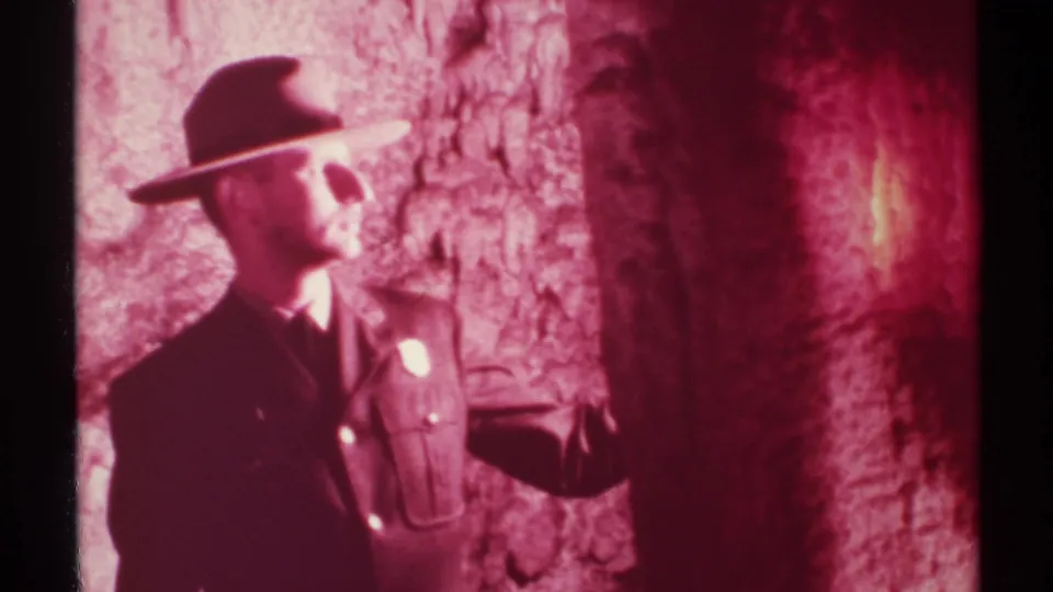 1972 New Mexico 8mm frame of a hat-wearing uniformed adult looking upward inside a cave as a flashlight lights the rock.