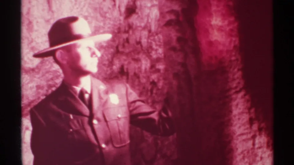 1972 New Mexico 8mm frame showing a uniformed man in profile beside a reddish cave wall, with his arm raised near the rock.