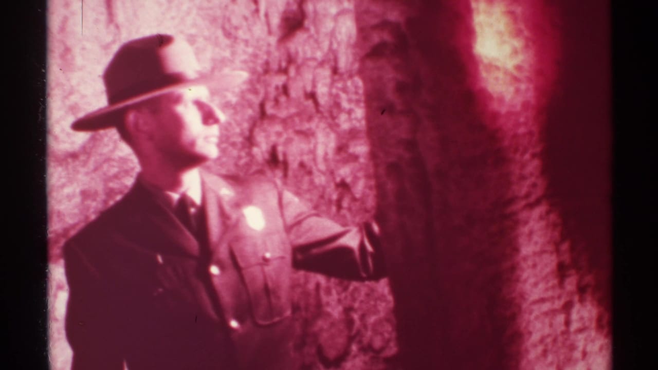 Uniformed ranger in a wide-brim hat stands beside a red-lit cave wall.