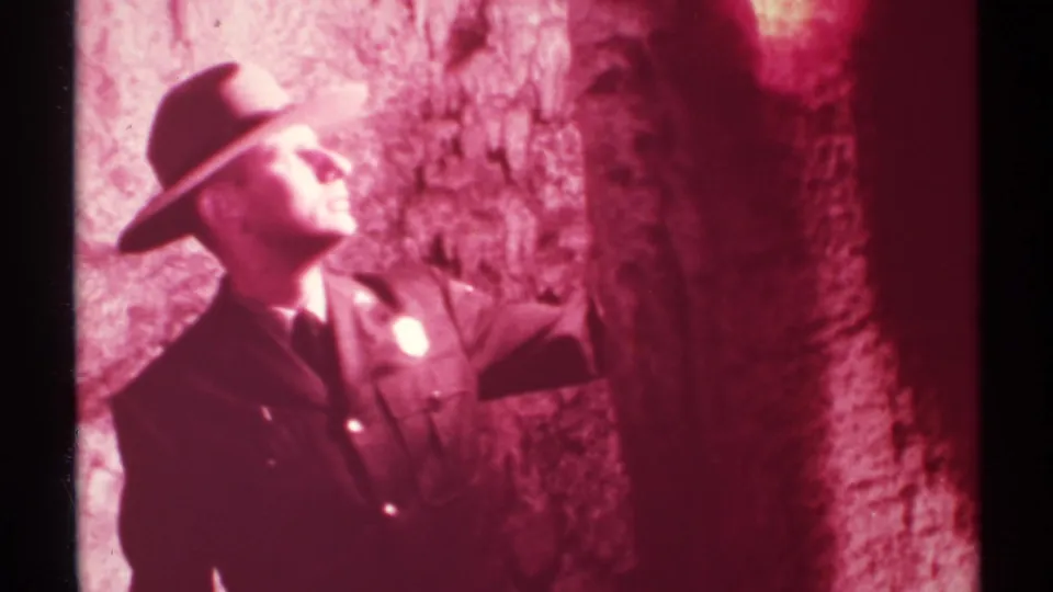 Uniformed ranger in a New Mexico cave appears to gesture toward red-lit rock formations in a 1972 8mm frame.