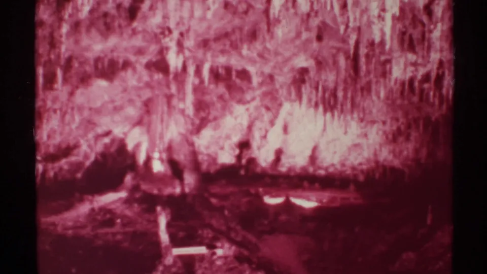 1972 8mm New Mexico cave footage showing red-magenta stalactites, shadowed rock, and faint reflected light in a low cave chamber.