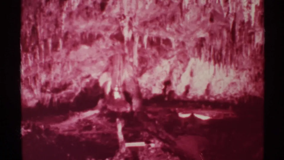 1972 8mm New Mexico cave interior with dense stalactites, red color cast, dark lower rock, and small reflected highlights.