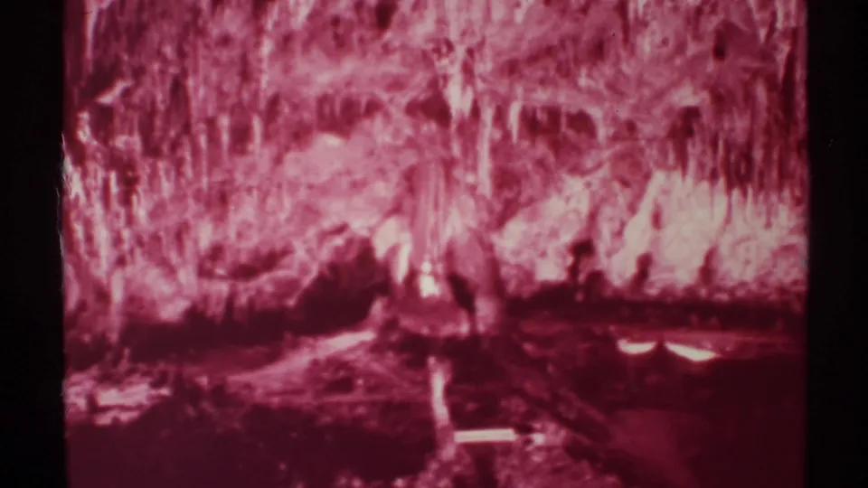 1972 8mm New Mexico cave shot showing blurred red-lit formations, a dark pool or floor area, and a pale ledge-like line in the foreground.