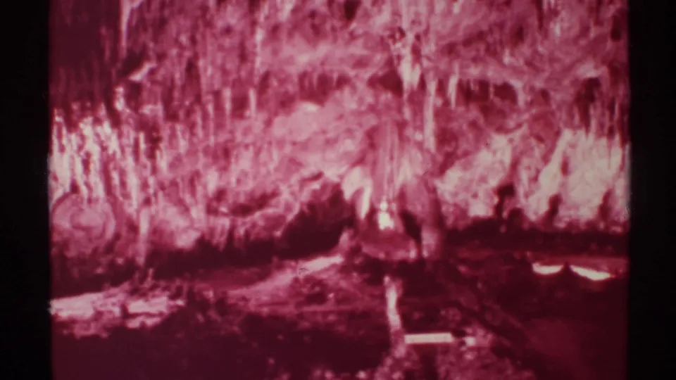 1972 8mm New Mexico cave interior with red-magenta stalactites, rough wall texture, dark lower chamber, and reflected light.