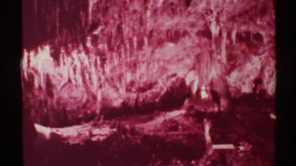 1972 New Mexico 8mm frame of a cave interior in strong pink cast, showing vertical formations, shadowed floor, and a small bright marker-like shape.