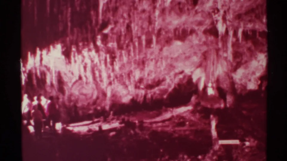 1972 New Mexico home-movie frame of a red-tinted cave chamber with hanging formations and small visitor figures at the edge.