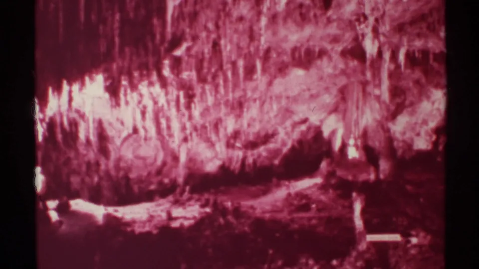 1972 New Mexico cave footage frame with red-tinted formations, a dark walkway area, and small visitor figures near the lower left.