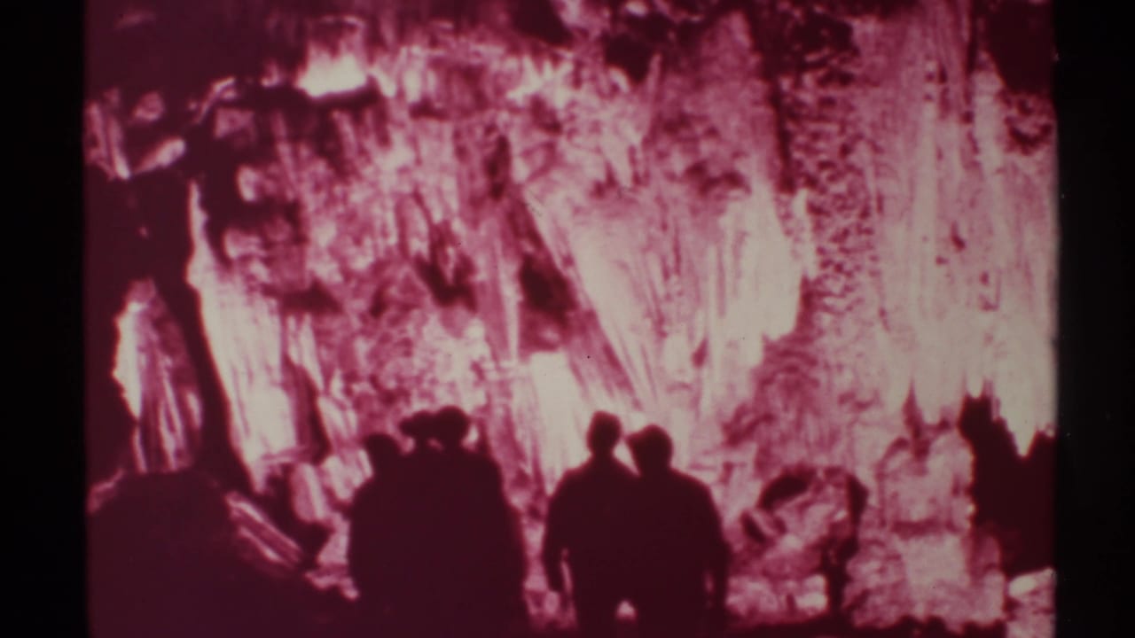 Silhouetted visitors stand before a pink-lit cave wall with bright rock formations.