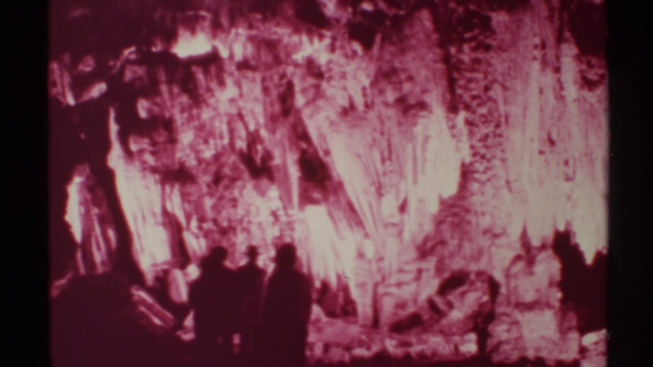A small group walks through a pink-lit cavern with dark figures in the foreground.