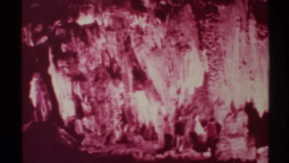 A handheld 8mm view shows men near the lower edge of a red-tinted cavern scene, with textured rock formations filling the frame.