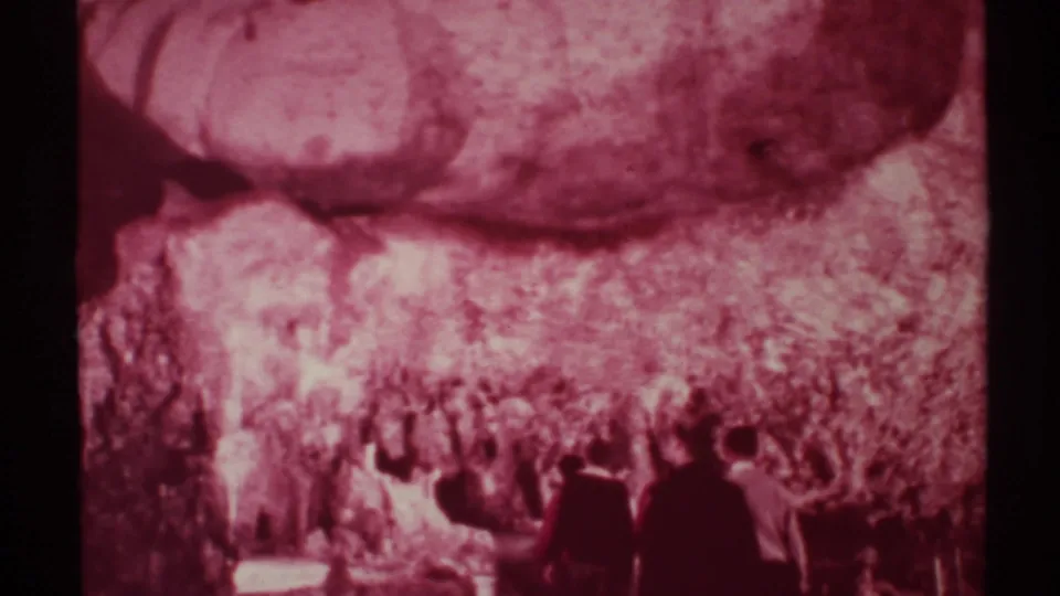 Red-tinted 1972 New Mexico home-movie frame of formally dressed adults walking into a cavern interior.
