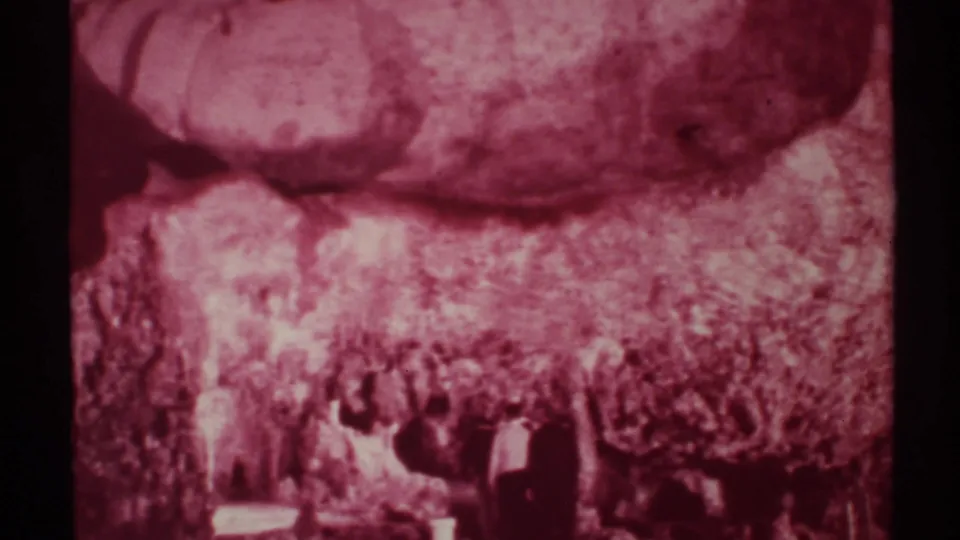 1972 New Mexico cave footage frame with distant people near the lower edge and rough stone formations filling the view.