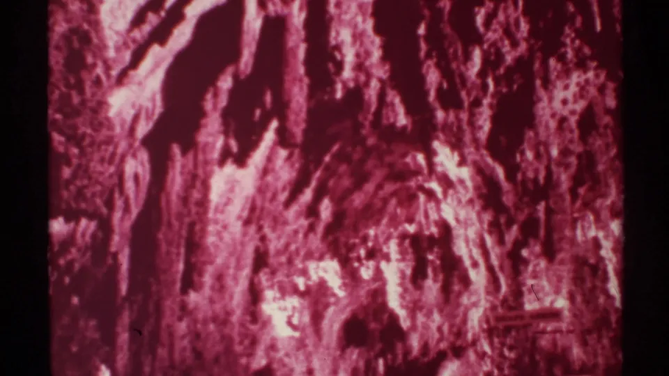 1972 8mm New Mexico cave view with jagged pink-red formations, shadowed openings, and a small faint sign-like detail near the lower right.