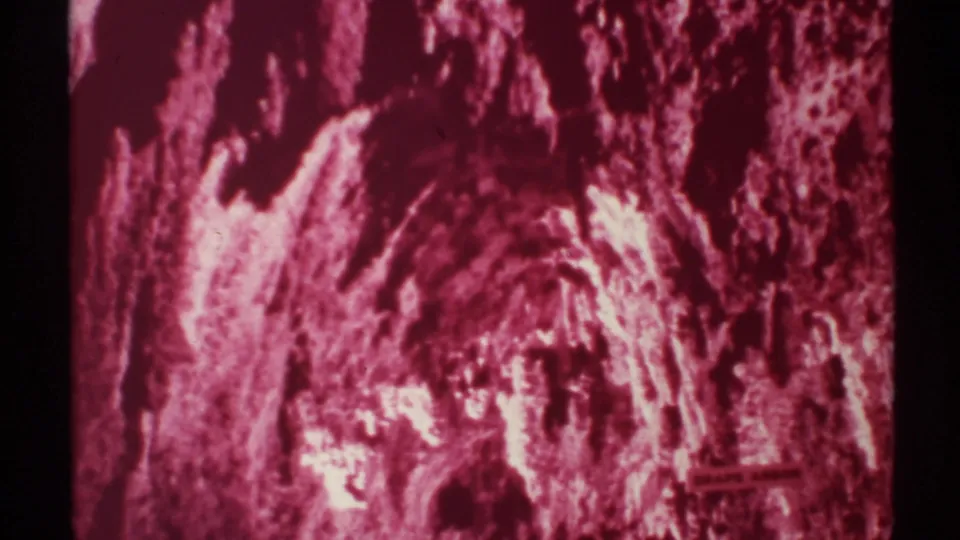 1972 8mm New Mexico cave interior with rough pink-red formations, heavy shadow, and a small lower-right sign or marking partly visible.