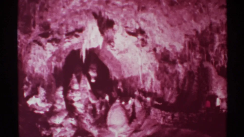 1972 New Mexico cave footage frame with soft focus, magenta tint, and a dark recess beneath rough stone formations.