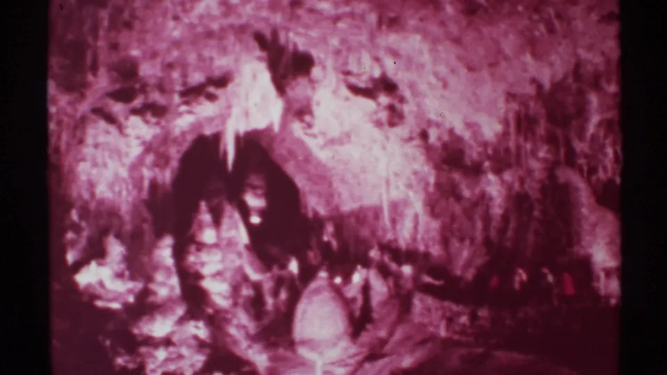 1972 New Mexico cave frame with rugged stone formations and faint visitor figures in the dim lower area.