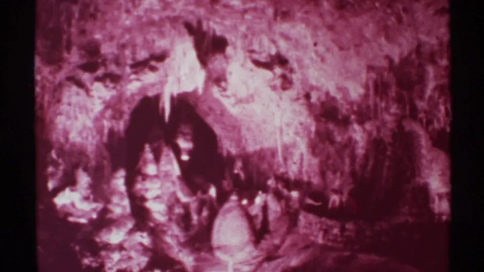 Aged 8mm frame of a dim cavern walkway in New Mexico, with people barely visible against textured rock and pale formations.