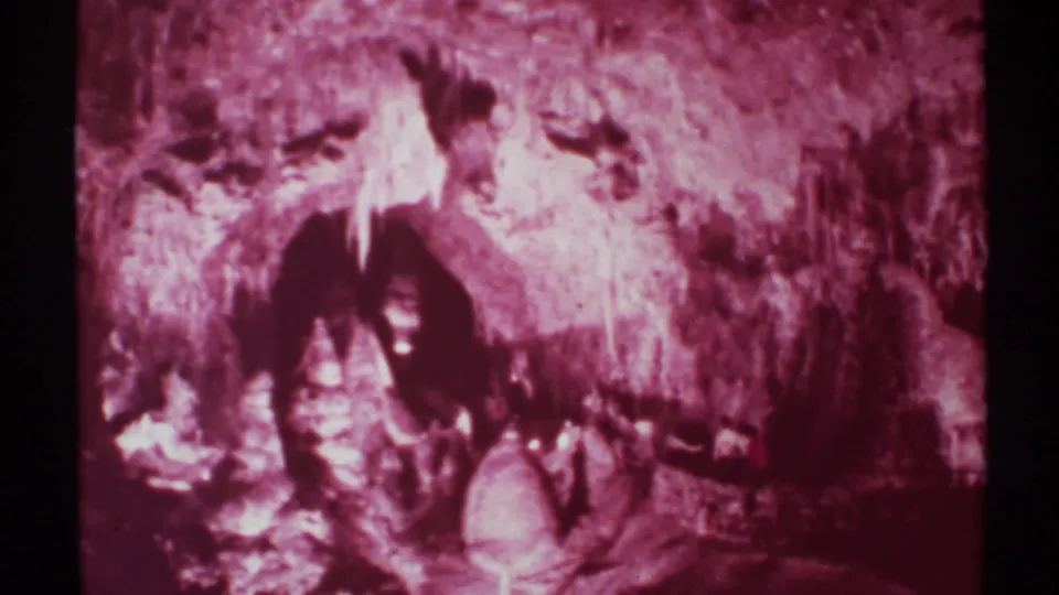 Soft 1972 8mm frame of a New Mexico cave interior, showing reddish rock texture and a partly visible person in a dark recess.