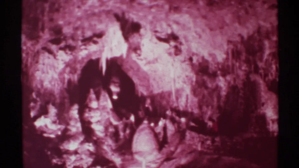 Grainy 1972 8mm New Mexico cave frame with jagged walls, bright mineral-like streaks, and a person partly hidden by shadow.