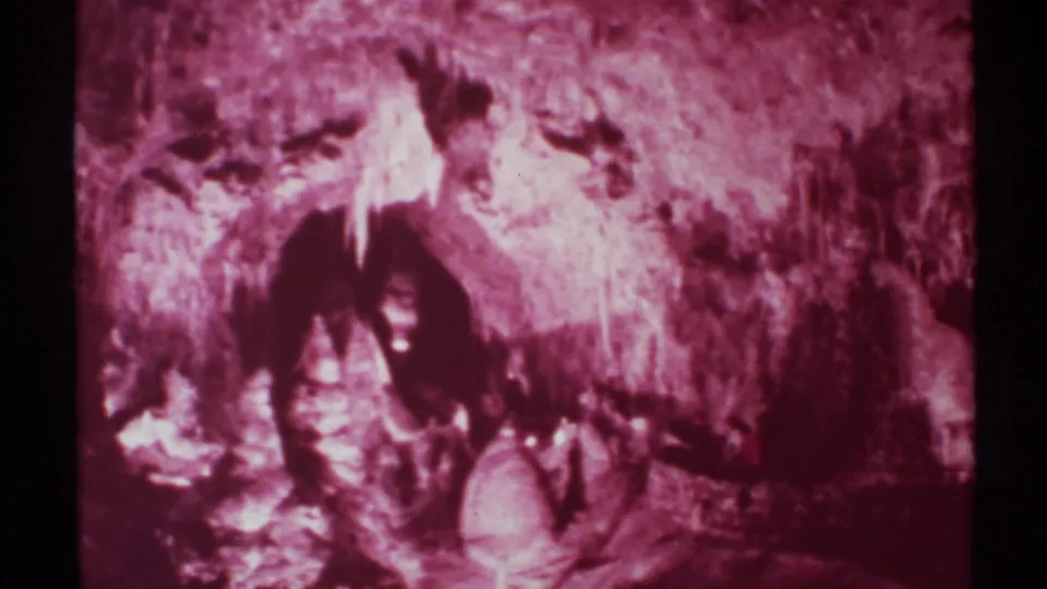 Final 1972 8mm New Mexico cave view with heavy maroon color shift, rough stone surfaces, and a barely defined person in the center.