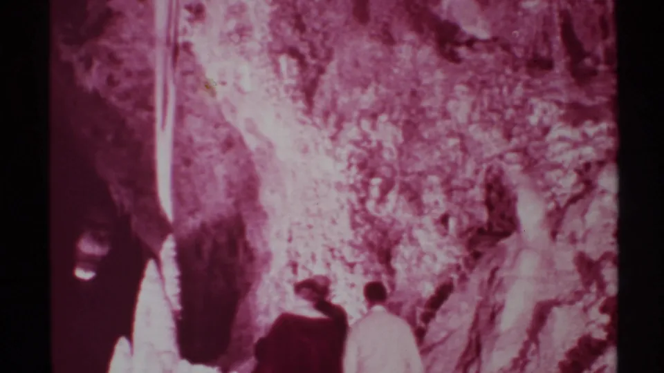 A handheld 1972 New Mexico frame shows cave visitors from behind moving past illuminated formations and shadowed stone.