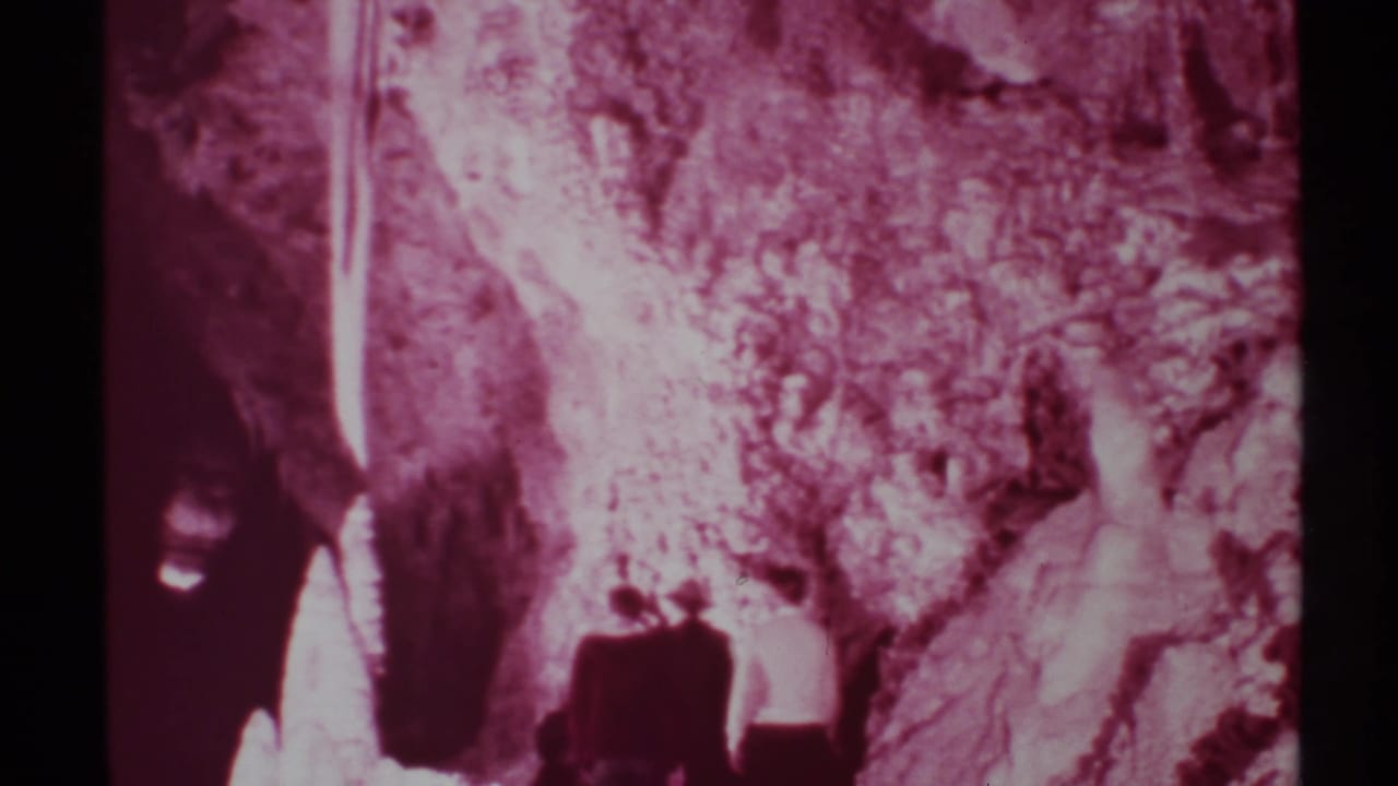 1972 New Mexico 8mm frame showing small visitors near a cave entrance with maroon tint, shadows, and textured rock walls.