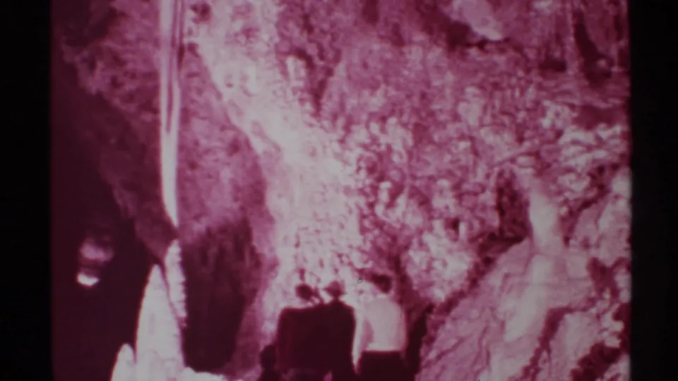 1972 New Mexico 8mm frame showing small visitors near a cave entrance with maroon tint, shadows, and textured rock walls.