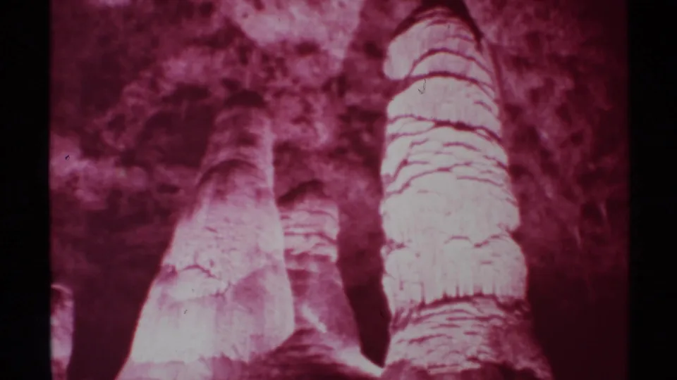 1972 New Mexico home-movie frame focused on pale stalagmite-like cave formations, rough rock texture, and heavy maroon coloration.