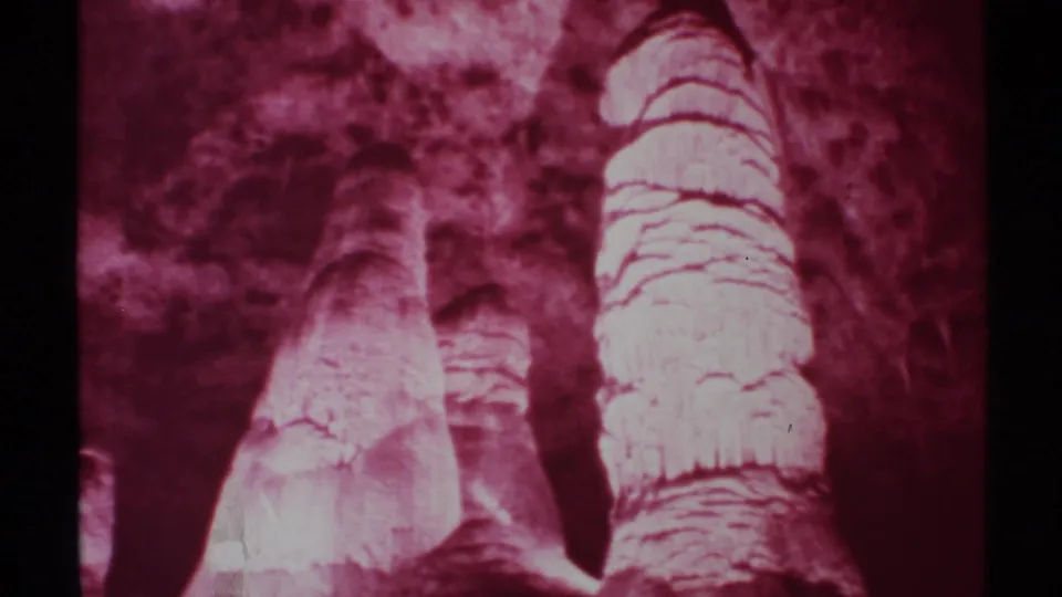 Maroon-tinted 8mm frame from a New Mexico cave visit showing two tall pale formations and shadowed rock surfaces.