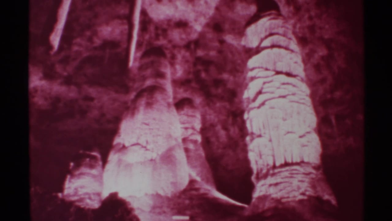 1972 8mm New Mexico cave interior showing red-tinted stalagmites, stalactites, layered mineral surfaces, and dark surrounding rock.