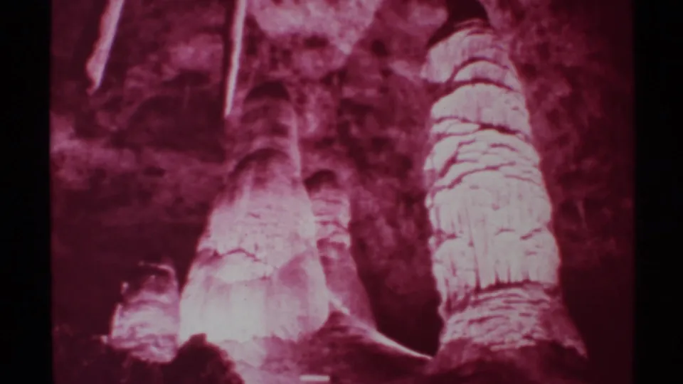 1972 8mm New Mexico cave interior showing red-tinted stalagmites, stalactites, layered mineral surfaces, and dark surrounding rock.