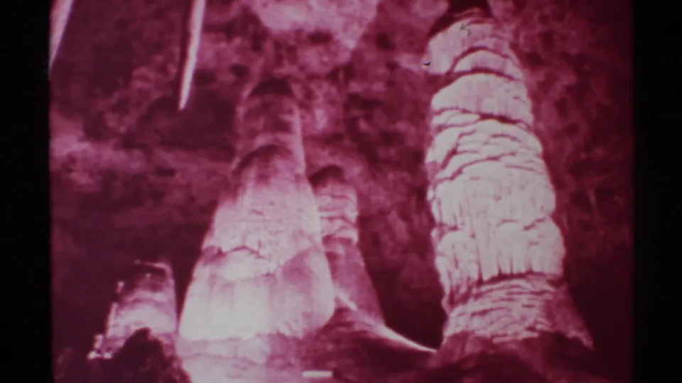 1972 8mm New Mexico cave view with soft red exposure, clustered stalagmites, hanging formations, and textured rock walls.