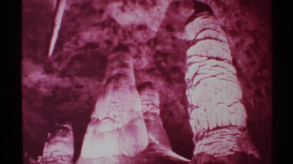 1972 New Mexico cave footage frame with red-tinted limestone formations, including a large stalagmite cluster and dark surrounding rock.