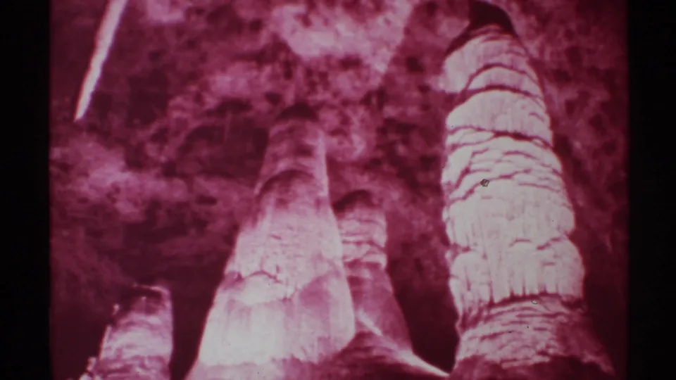 1972 New Mexico 8mm frame showing a tall red-lit stalagmite beside smaller cave formations and shadowed cavern walls.