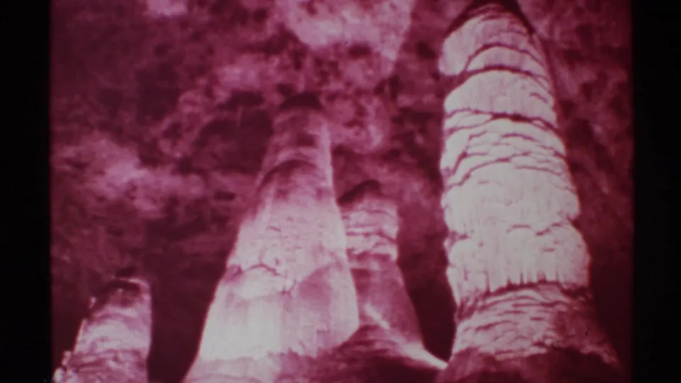 1972 New Mexico 8mm frame showing red-filtered cave formations, including tall pale rock columns against a darker cavern wall.