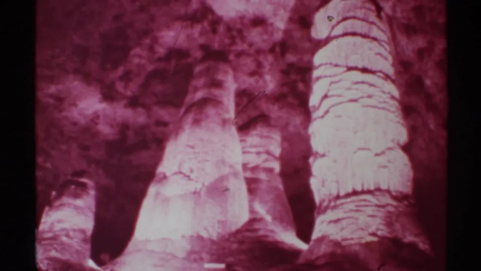 1972 8mm frame of red-toned cave formations in New Mexico, with a large pale column, smaller columns, and granular film texture.