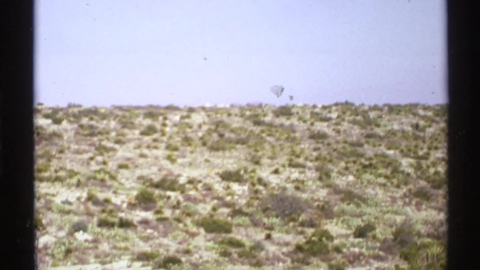 1972 8mm frame of a dry New Mexico landscape with low scrub, rocky ground, and a pale open sky.