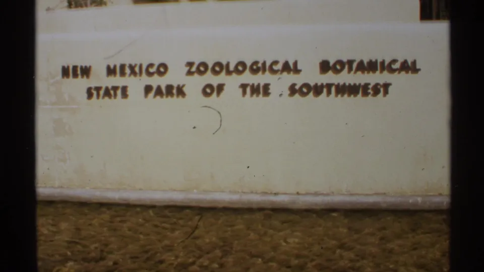The 1972 home-movie frame holds on the New Mexico park sign, with raised letters, cream surface, and bare soil below.