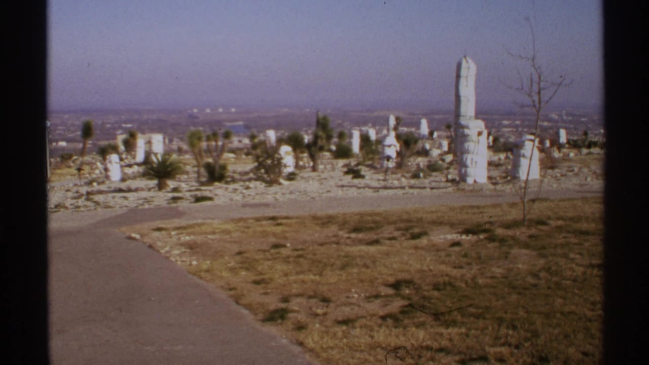 White stone monoliths on a dry hillside beside a paved path with a hazy city beyond.