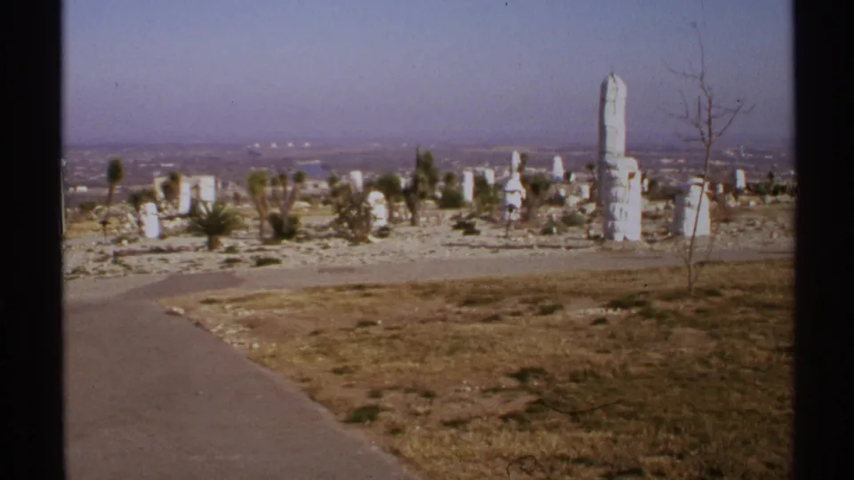 1972 New Mexico 8mm frame showing tall white stone forms, desert plants, dry grass, and a distant city under haze.