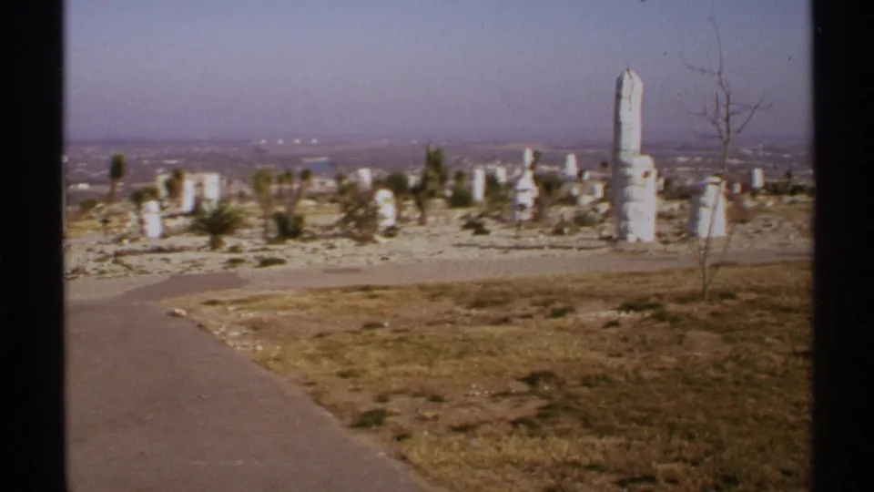 1972 New Mexico home-movie frame of a curved walkway, white stone columns, sparse plants, and a hazy skyline.