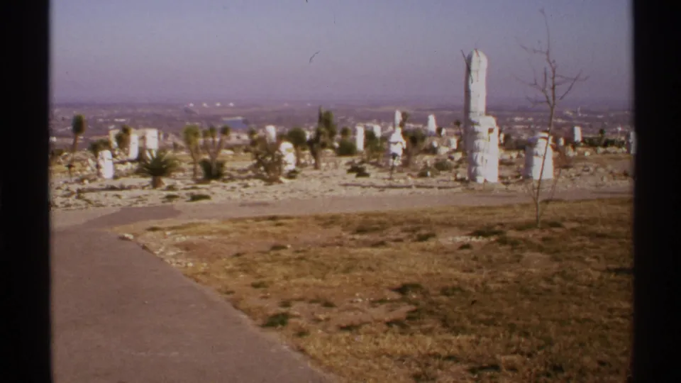 1972 New Mexico 8mm view of white sculptural stones, a paved path, dry ground, sparse plants, and city haze.