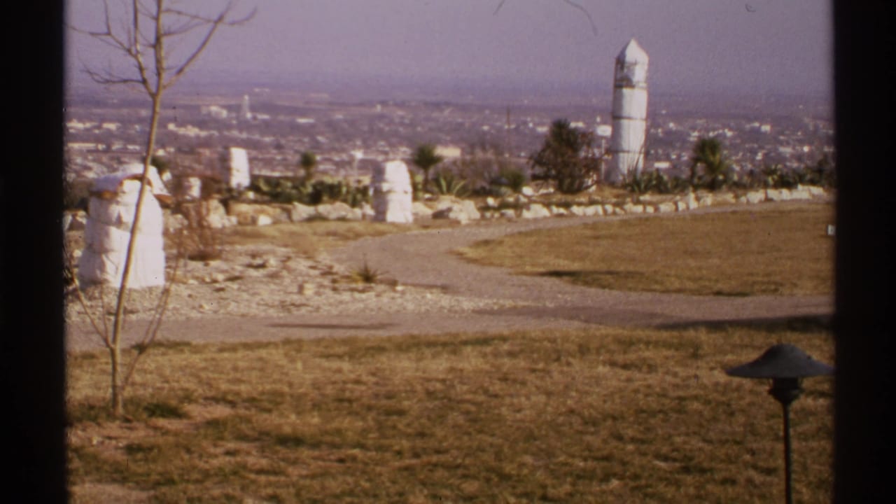 1972 New Mexico home-movie view of a dry zoo-like overlook with a curved path, bare tree, white structures, and distant city haze.
