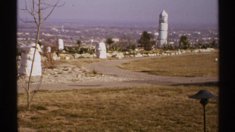 1972 New Mexico home-movie frame of a sunlit dry overlook, curved path, sparse vegetation, and hazy city in the distance.