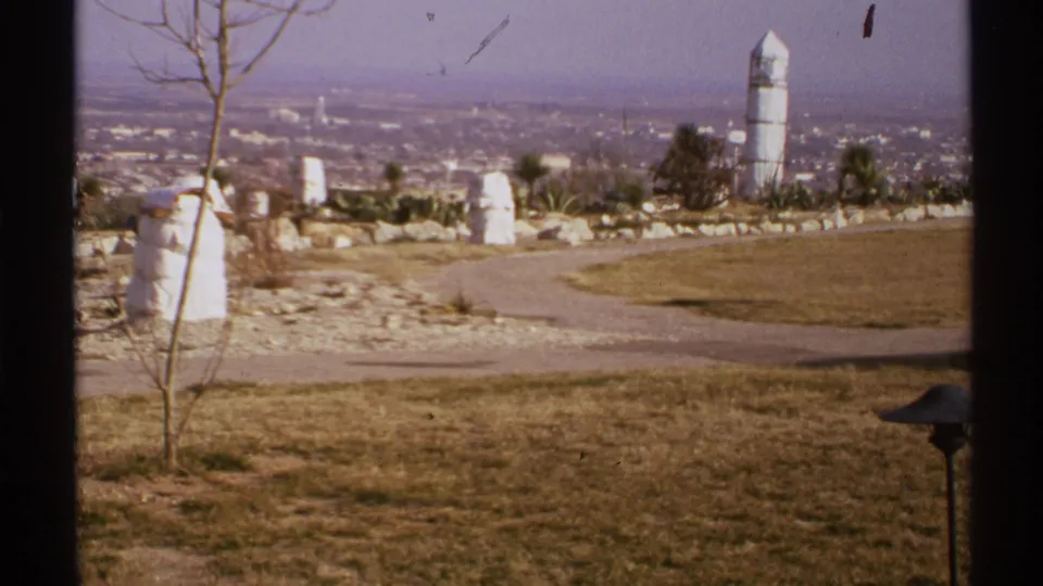 1972 New Mexico home-movie frame showing a dry zoo-like landscape with a curved path, sparse plants, white upright forms, and city haze beyond.