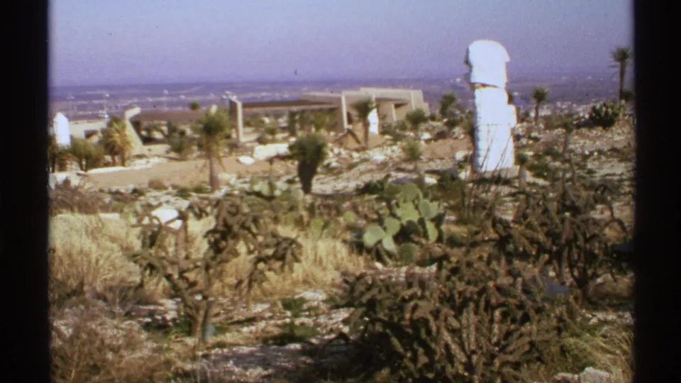 1972 New Mexico 8mm frame of a dry desert landscape with cactus, palms, pale structures, and a hazy valley beyond.
