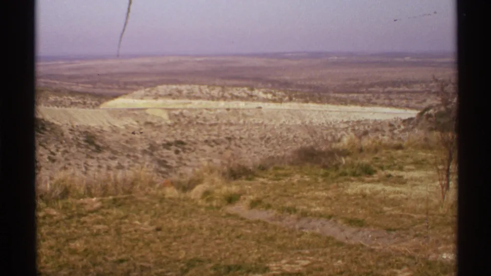 1972 New Mexico 8mm landscape frame showing a broad arid plain, pale road or wash, scrub vegetation, and hazy purple sky from an overlook.